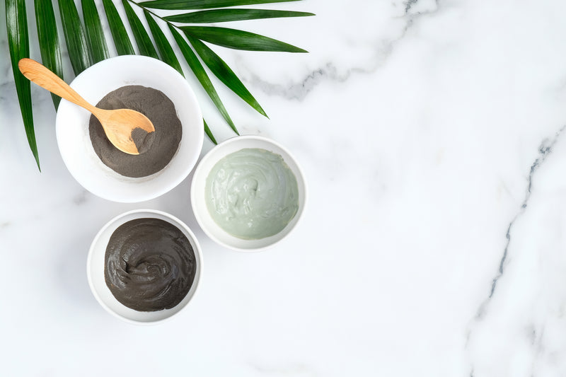 Activated charcoal powder and Dead Sea Mud mask in two separate white bowls on marble table